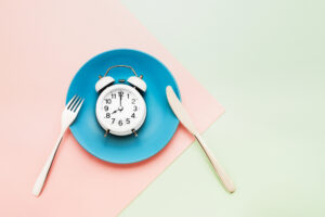 Intermittent fasting concept. White alarm clock on empty blue dish with knife and fork on pink-green background. top view, copy space for text.